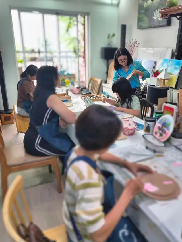 Học viên nhiều độ tuổi cùng vẽ và làm đồ thủ công quanh bàn lớn trong một buổi workshop